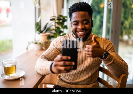 Ein junger schwarzer Mann sitzt in lässiger Kleidung an der Bar. Er nutzt ein Smartphone und führt Videoanrufe. Stockfoto