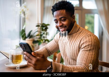 Porträt eines glücklichen schwarzen Mannes, der an der Bar sitzt, lässig gekleidet. Er nutzt Smartphone und Messaging online. Stockfoto