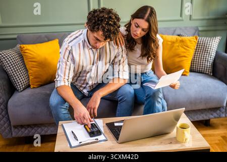 Ein paar, das über Hausfinanzen spricht, während er Rechnungen prüft. Sie überprüfen die Finanzdokumente und berechnen das Familienbudget im Wohnzimmer. Stockfoto
