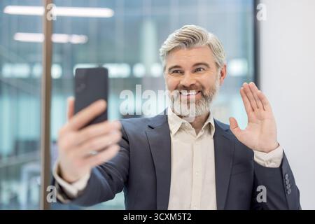 Reifer Geschäftsmann lächelt und winkt während eines Videoanrufs mit dem Smartphone in einem modernen Büro, begrüßt Kollegen oder Kunden und engagiert sich für freundlich, prof Stockfoto