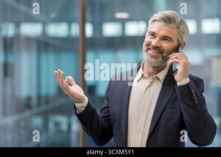 Reifer Geschäftsmann, der während eines Telefonats lächelnd und gespenstisch in einem Büro mit moderner Architektur steht und erfolgreiche Kommunikation und Kommunikation repräsentiert Stockfoto