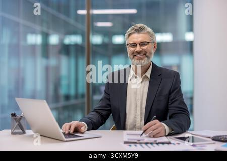 Glücklicher, reifer Geschäftsmann, der an einem Laptop arbeitet und Dokumente an einem Schreibtisch in einem modernen Büro liest, steht für Erfolg, Professionalität und Erfahrung Stockfoto