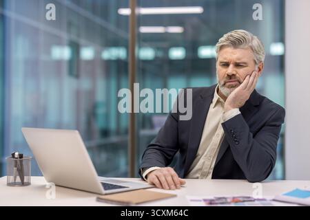 Reifer erfahrener Geschäftsmann, der am Arbeitsplatz krank ist, an Zahnschmerzen leidet und Hand an Gesicht hält Stockfoto