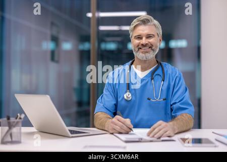 Ein lächelnder männlicher Arzt trägt blaue Peelings und Stethoskop, sitzt an einem Schreibtisch, während er auf ein Klemmbrett schreibt, repräsentiert Professionalität und engagierte ihn Stockfoto