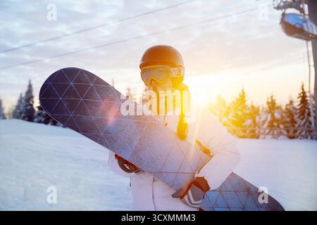 Porträt der Frau auf dem Hintergrund von Bergen und Sonnenuntergang. Ein Snowboard halten und Skibrille tragen Stockfoto