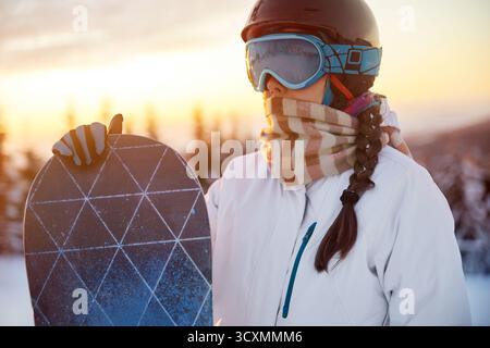 Porträt der Frau auf dem Hintergrund von Bergen und Sonnenuntergang. Ein Snowboard halten und Skibrille tragen. Skibrille Des Snowboarders Mit Der Reflecti Stockfoto