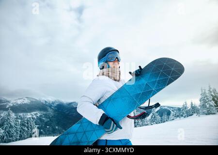 Porträt der Frau auf dem Hintergrund von Bergen und Sonnenuntergang. Ein Snowboard halten und Skibrille tragen. Skibrille Des Snowboarders Mit Der Reflecti Stockfoto