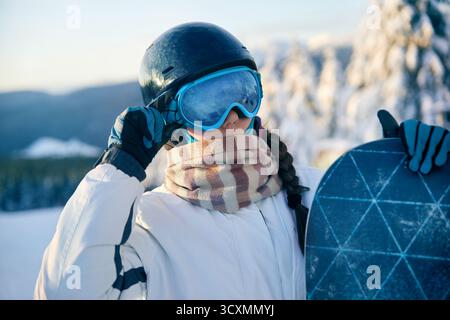Porträt einer Frau, die ein Snowboard hält und Skibrille trägt. Skibrille Von Snowboardern Mit Dem Spiegelbild Der Verschneiten Berge. Stockfoto