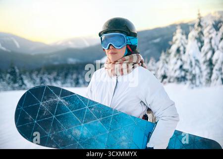 Porträt der Frau auf dem Hintergrund von Bergen und Sonnenuntergang. Ein Snowboard halten und Skibrille tragen. Skibrille Des Snowboarders Mit Der Reflecti Stockfoto