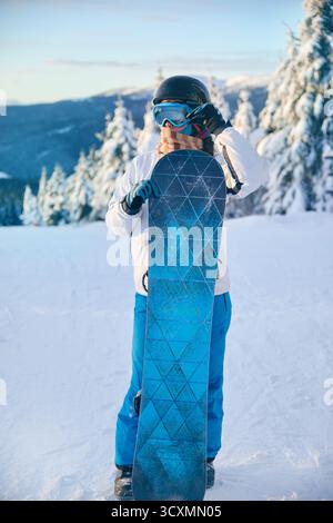 Porträt einer Frau, die ein Snowboard hält und Skibrille trägt. Skibrille Von Snowboardern Mit Dem Spiegelbild Der Verschneiten Berge. Stockfoto