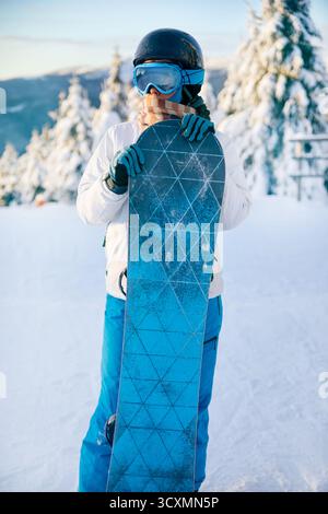 Porträt einer Frau, die ein Snowboard hält und Skibrille trägt. Skibrille Von Snowboardern Mit Dem Spiegelbild Der Verschneiten Berge. Wintersport Stockfoto