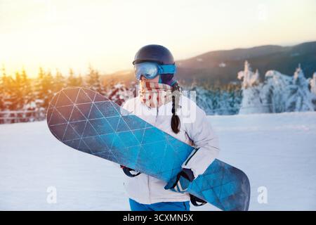 Porträt der Frau auf dem Hintergrund von Bergen und Sonnenuntergang. Ein Snowboard halten und Skibrille tragen. Skibrille Des Snowboarders Mit Der Reflecti Stockfoto