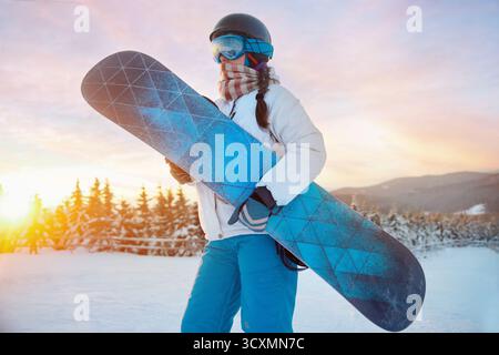 Porträt der Frau auf dem Hintergrund von Bergen und Sonnenuntergang. Ein Snowboard halten und Skibrille tragen. Skibrille Des Snowboarders Mit Der Reflecti Stockfoto