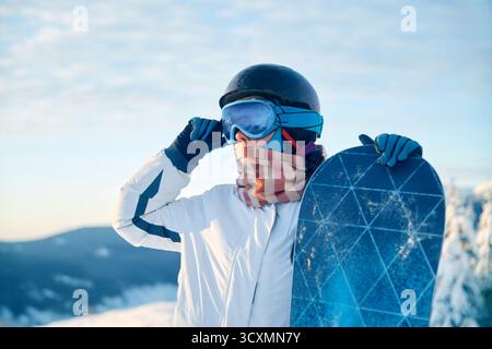 Porträt einer Frau, die ein Snowboard hält und Skibrille trägt. Skibrille Von Snowboardern Mit Dem Spiegelbild Der Verschneiten Berge. Wintersport Stockfoto