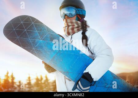 Porträt der Frau auf dem Hintergrund von Bergen und Sonnenuntergang. Ein Snowboard halten und Skibrille tragen. Skibrille Des Snowboarders Mit Der Reflecti Stockfoto