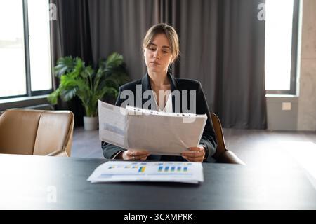 Geschäftsfrau in einem formellen schwarzen Anzug, die an einem Schreibtisch sitzt und die Finanzdokumente und Berichte des Unternehmens sorgfältig überprüft und analysiert Stockfoto