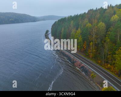 Weite Sicht aus der Luft auf eine lange, leere Asphaltstraße am Ufer eines breiten, dunklen Sees, mit einem dichten Wald mit farbenfrohen Herbstlaub Stockfoto