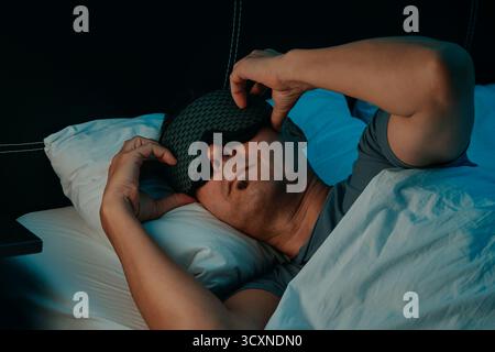 Nahaufnahme eines Mannes im Bett, der eine gemusterte Schlafmaske mit beiden Händen in einem Schlafzimmer anpasst, auf einem Kissen unter einem weißen Bettlaken liegend, in einem grauen T shi Stockfoto
