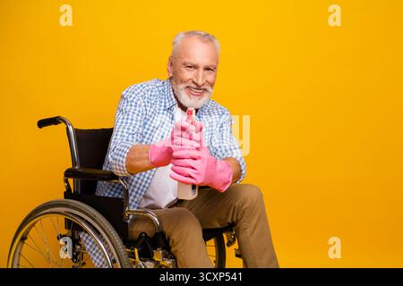 Lustiger herzerwärmender Opa im Rollstuhl lächelt, während er eine Sprühflasche auf hellgelbem Hintergrund benutzt, was Inklusionspflege und Reha-Erfolg zeigt Stockfoto