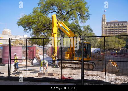 Die Crew-Mitglieder arbeiten auf einer umzäunten Baustelle mit einem gelben Komatsu-Bagger in der Nähe des Alamo Plaza, während die texanische Flagge auf einem nahe gelegenen Gebäude schwingt Stockfoto