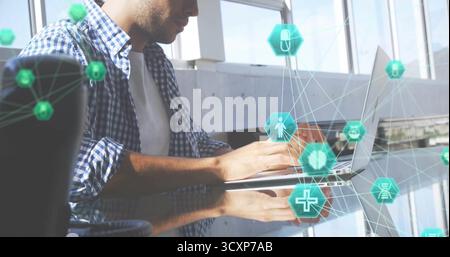 Ein hispanischer Mann in kariertem Hemd am Glasschreibtisch, mit silbernem Laptop und schwebenden digitalen Ikonen Stockfoto