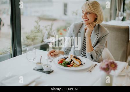 Eine stilvolle Frau sitzt an einem weiß gekleideten Tisch in einem modernen Restaurant. Sie lächelt, während sie ein Gourmet-Essen und ein Glas Wein bei Sonnenlicht genießt Stockfoto