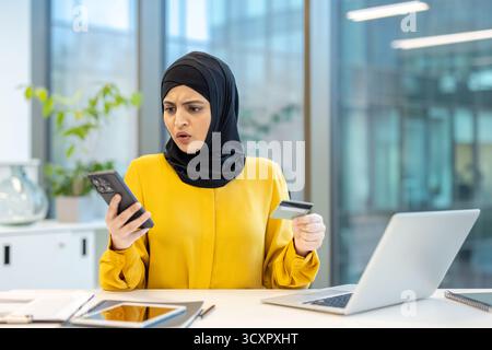 Muslimische Geschäftsfrau in einem Hijab, die Schock und Frustration ausdrückt, während sie auf ihr Smartphone blickt und eine Kreditkarte hält und dabei ein Onlin erlebt Stockfoto