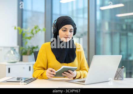 Junge ernsthafte muslimische Frau mit schwarzem Hijab und Kopfhörern, sitzt an einem modernen Schreibtisch, studiert online und macht sich Notizen auf einem Notizblock Stockfoto