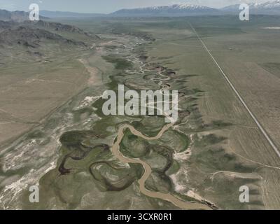 Der sich windende Wasserweg schafft natürliche Muster durch eine grüne Ebene mit weit entfernten Bergen und einer langen geraden Straße, die sich über die ausgedehnte Landschaft erstreckt Stockfoto
