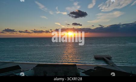 Sonnenaufgang am Horizont über der ruhigen Meeresoberfläche, das goldene Licht reflektiert auf den Sandstrand und einen hölzernen Pier, während sich Wolken am Himmel sammeln Stockfoto