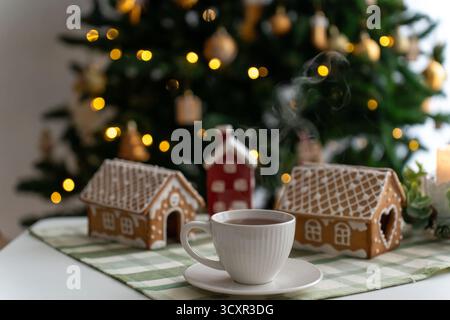 Gemütliche Tasse heißer Tee auf dem Tisch in der Nähe von Lebkuchenhäusern und Weihnachtsbaum mit goldenen Lichtern. Konzept von Winterferien, Wohnkomfort, festliche Stimmung, tr Stockfoto