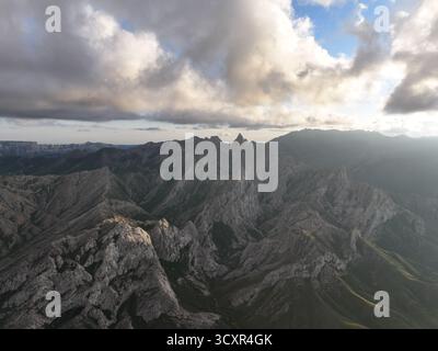 Zerklüftete Bergketten durchqueren eine weite Landschaft unter einem dynamischen Himmel voller dunkler Wolken und blauer Flecken, die Konzepte von Natur, Macht, Stockfoto