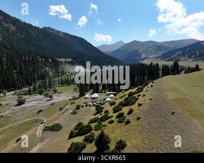 Jurtencamp mit traditionellen Wohnhäusern und Fahrzeugen in einem grünen Tal, umgeben von hoch aufragenden Kiefernwäldern unter einem hellblauen Himmel, bietet CUL Stockfoto