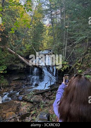 Der Rücken einer Frau, die ein Foto von Wagner Falls macht, die über Felsen und umgestürzte Bäume einen Hügel hinunterfallen, im Wald in Munising, Michigan, USA Stockfoto