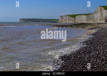 Seven Sisters Cliffs, die sich vom Ärmelkanal erheben, bieten einen unberührten Kiesstrand und sanfte Wellen unter einem klaren blauen Himmel. East Sussex, England Stockfoto