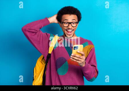 Ein glücklicher junger Mann mit pulsierendem Pullover überprüft das Telefon, während er den Rucksack vor hellblauem Hintergrund hält Stockfoto