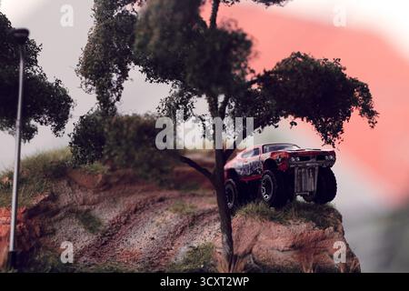 Rot-schwarz Muscle Car steht kühn auf einem felsigen Hügel, umgeben von strukturiertem Boden, Gras und Bäumen, die an eine wilde Landschaft im Freien erinnern. Die Szene Stockfoto