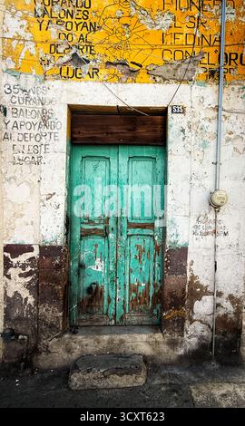 Eine verwitterte türkisfarbene Holztür mit abblätternder Farbe in einer alten Wand, die mit verblassten Werbespots und Graffiti bedeckt ist. Stockfoto