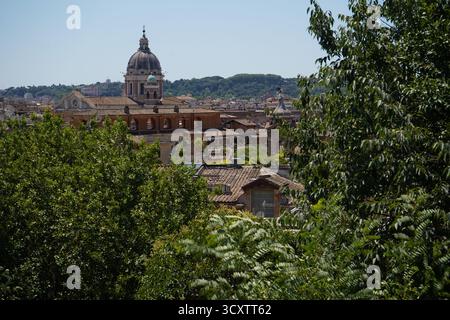 Blick aus nächster Nähe, eingerahmt von üppigen Bäumen, die die Terrakotta-Dächer hervorheben, eine große prominente barocke Kirchenkuppel, die die römische Architektur dominiert. Stockfoto