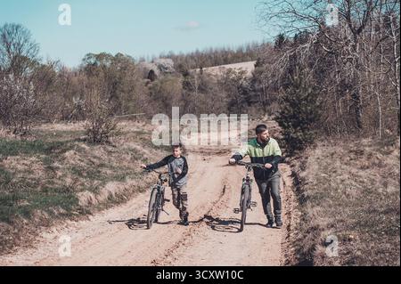 Ein Mann und ein Junge laufen mit ihren Fahrrädern auf einem unbefestigten Weg. Bäume und eine hügelige Landschaft bilden den Hintergrund. Sie scheinen eine gemächliche Outdoor-Show zu genießen Stockfoto
