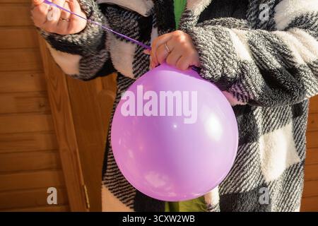 Junge Frau im warmen Mantel mit rosa Ballon drinnen in der Nähe der sonnigen Glastür mit Blick auf den Garten. Stockfoto