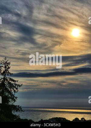 Meereslandschaft bei Sonnenuntergang oder Sonnenaufgang. Der Himmel ist mit langen horizontalen Wolken in Blau-, Grau- und Hellgelb bedeckt. Die Sonne scheint eine helle Scheibe durch Stockfoto