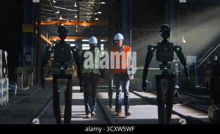 Ingenieure für Sicherheitswesten und Helme arbeiten mit humanoiden Robotern in der High-Tech-Industrieanlage zusammen. Zukunft der intelligenten Fertigung nahtlose Teamwork von Human Robot, Innovation in der Produktion. Stockfoto