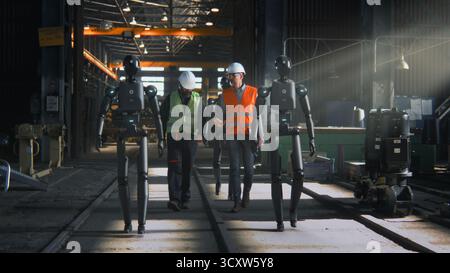 Männliche Ingenieure, die Schutzwesten und Helme tragen, arbeiten zusammen mit humanoiden Robotern bei Large Industrial Enterprise. Konzept der Fertigung der Zukunft, Zusammenarbeit zwischen Mensch und Roboter. Stockfoto