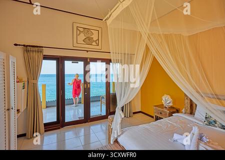 Eine Frau in rotem Kleid genießt den Meerblick auf der Terrasse ihres Hotelzimmers mit Himmelbett. Amed, Karangasem Regency, Bali, Indonesien. Stockfoto