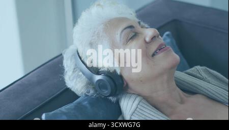 Liegende ältere Frau mit Strickjacke und Kopfhörern auf der Wohnzimmercouch mit blauem Kissen Stockfoto