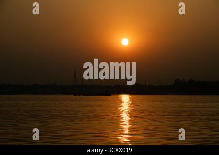 Eine blendende goldene Sonne und ihr Spiegelbild dominieren die Flussszene, die ein warmes, helles Leuchten auf das dunkle Profil des fernen Ufers wirft Stockfoto