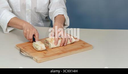 Appetitliche Shawarma wird mit einem Messer auf Holzoberfläche geschnitten. Frau in weißem Hemd schneidet Rollen mit Fleisch und Gemüse. wir kochen Fastfood zu Hause. Stockfoto