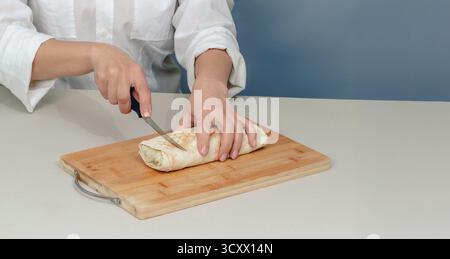 Appetitliche Shawarma wird mit einem Messer auf Holzoberfläche geschnitten. Frau in weißem Hemd schneidet Rollen mit Fleisch und Gemüse. wir kochen Fastfood zu Hause. Stockfoto