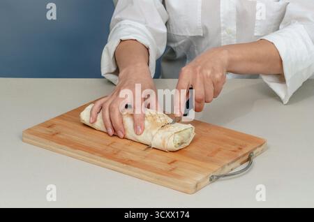 Appetitliche Shawarma wird mit einem Messer auf Holzoberfläche geschnitten. Frau in weißem Hemd schneidet Rollen mit Fleisch und Gemüse. wir kochen Fastfood zu Hause. Stockfoto